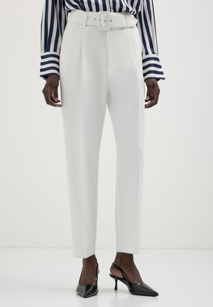 Pantalon blanc taille haute sur mesure avec ceinture, porté avec des escarpins slingback noirs à bout pointu et une chemise à manches longues rayée noire et blanche.