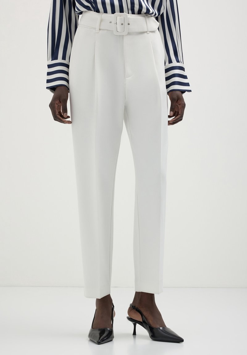 Pantalon blanc taille haute sur mesure avec ceinture, porté avec des escarpins slingback noirs à bout pointu et une chemise à manches longues rayée noire et blanche.
