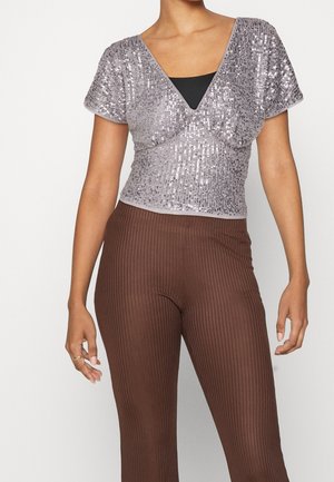 Person trägt ein kurzärmeliges silbernes Paillettenoberteil über einem schwarzen Unterhemd und hoch taillierte, gerippte braune Hosen, steht vor einem weißen Hintergrund.