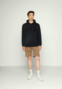 Joven de pie con una sudadera negra de GAP, pantalones cortos beige, calcetines blancos y zapatillas blancas frente a un fondo claro y simple.