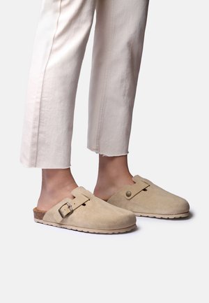 Stopy w beżowych wsuwanych chodakach zamszowych z klamrami, w połączeniu z jasnymi spodniami 7/8, stojące na prostym tle.