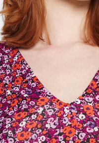 Tričko s výstřihem do V, zdobené multibarevným květinovým vzorem s fialovými, oranžovými a bílými květy na tmavém pozadí; jemná pletená textura.