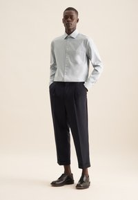 Hellblaues Langarmhemd mit einer Tasche, kombiniert mit schwarzen, gefalteten Hosen und schwarzen Lederschuhen. Glattes Material mit einem festen Kragen.
