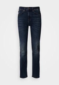 Σκούρο μπλε τζιν με ίσια γραμμή, με παραδοσιακό σχέδιο πέντε τσεπών, ορατές ραφές και κλείσιμο με κουμπί στη μέση.