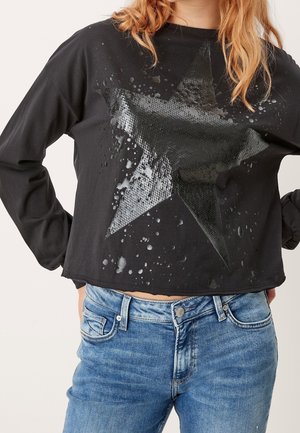 Sort langærmet cropped skjorte med stor tekstureret stjerne og malingstænk design, båret med blå jeans, hænderne på hofterne.