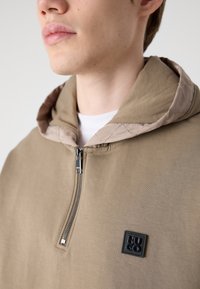 Sudadera marrón con cremallera frontal, capucha acolchada en beige y un parche de logo negro. La tela es suave y tiene un corte relajado.