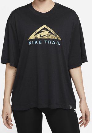 Czarna oversize koszulka Nike Trail z złotym trójkątnym logo i jasnoniebieskim napisem, noszona przez osobę z tatuażami na obu przedramionach.