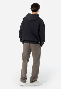 Sweatshirt preto com capuz, com um corte descontraído e punhos canelados, combinado com calças cinza claro. Modelo a usar sapatilhas brancas, visto de costas.