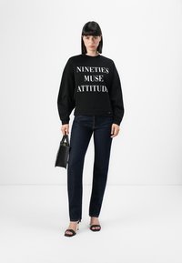 Felpa nera con testo bianco audace che recita "NINETIES MUSE ATTITUDE," abbinata a jeans scuri e tacchi neri, con una borsetta mini.