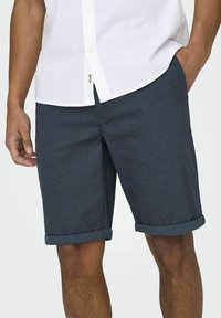 Shorts bleu foncé en tissu texturé, avec des ourlets roulés et une coupe décontractée. Associés à une chemise blanche à boutons.