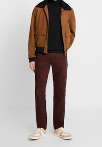 Giacca marrone in misto lana con colletto in pelliccia nera, abbinata a un dolcevita nero, pantaloni bordeaux e sneaker beige con strisce bianche.