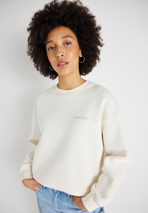 Sweatshirt de cor creme com um gola redonda, punhos canelados e um detalhe em texto branco. O modelo usa-o com jeans de ganga de lavagem clara.