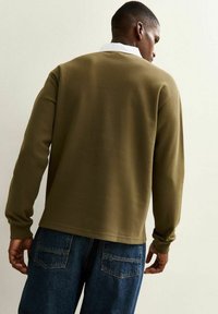 Olive green sweatshirt with a textured finish and long sleeves, paired with blue denim jeans, viewed from the back. White collar visible.