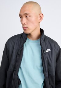 Κεφαλής άντρας με κρίκο σκουλαρίκι φοράει ανοιχτό μπλε πουκάμισο κάτω από μαύρη ζακέτα Nike με φερμουάρ, κοιτάζοντας στο πλάι σε απλό φόντο.