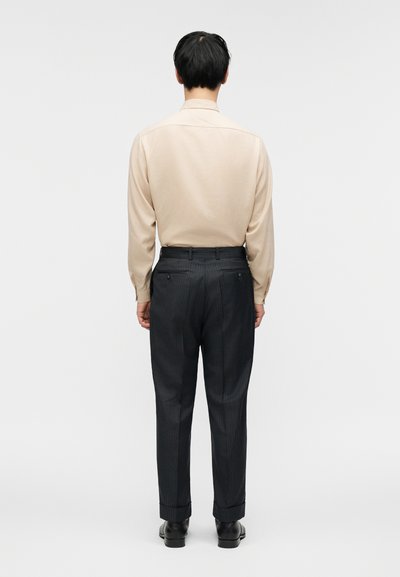 Άνδρας που στέκεται γυρισμένος από την πλάτη, φοράει μπεζ πουκάμισο με μακριά μανίκια, σκούρο παντελόνι με λεπτές ρίγες και μαύρα παπούτσια επίσημα, σε απλό φωτεινό φόντο.