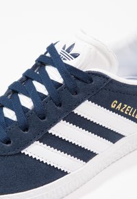 Blå semsket sneakers med hvite lærstriper, med marineblå snørebånd, teksturert såle og gull "Gazelle"-merke på siden.