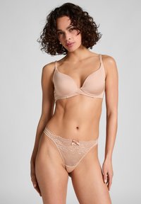 Beige gepolsterter BH mit einem Überkreuzdesign, verstellbaren Trägern, kombiniert mit einem passenden Spitzen-Thong, der eine Schleife und Seitenbänder hat.