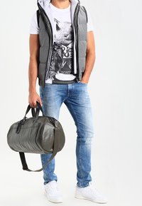 Grå pufferväst över en grafisk vit T-shirt, blå jeans och vita sneakers. Håller en grön läderväska med svarta handtag.