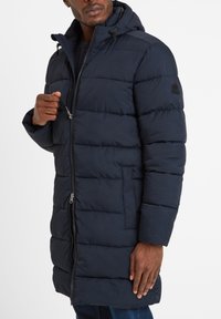 Man in dark blue padded coat with hood, partially zipped, hand holding zipper, standing against white background.