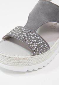 Sandale en daim gris avec une lanière texturée ornée de strass argentés et transparents, une plateforme blanche avec un détail tressé.