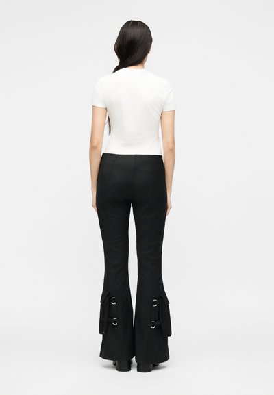 Γυναίκα που στέκεται με την πλάτη, φοράει λευκό κοντομάνικο τοπ και μαύρο παντελόνι καμπάνα με αγκράφες στις πλαϊνές τσέπες κοντά στους αστραγάλους.