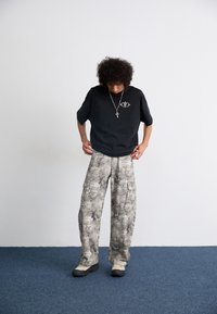 Black oversized t-shirt with front logo, paired with light gray patterned cargo pants featuring multiple pockets, and black and beige shoes.