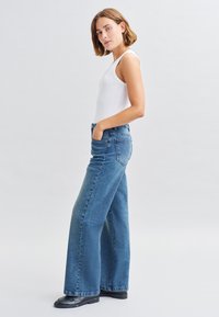 Jean en denim bleu à jambes larges et taille haute, associé à un débardeur ajusté blanc, mettant en avant des lignes épurées et un design classique.