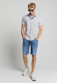 Korte mouw shirt met een witte basis en blauwe bladvormen, gecombineerd met denim shorts en grijze sneakers. Accessoires zijn onder meer een zonnebril en een blauwe sjaal.