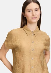 Camicia a maniche corte con colletto in tessuto dorato texture, caratterizzata da un motivo floreale. Chiusura con bottoni a contrasto di colore argento.