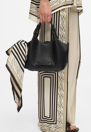 Roka drži črno teksturirano usnjeno torbico z zvezanimi ročaji in bež-črno črtasto vzorčeno rutko, oblečene v ujemajoče se vzorčaste hlače.