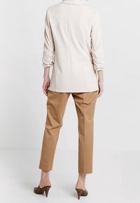 Světle béžový top s dlouhými rukávy a strukturovaným límcem, spárovaný s kalhotami v rovné siluetě v barvě tan. Tkanina vypadá hladce a precizně zpracovaně.