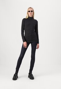 Zwarte lange mouwen turtleneck met een subtiel textuurpatroon, gecombineerd met donkere skinny jeans en chunky zwarte schoenen. Model poseert rechtop.