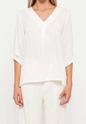 Femme portant un chemisier blanc ample à col en V avec manches trois-quarts et un pantalon blanc, debout devant un fond uni.