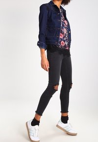 Džínová bunda, vzorované tričko s květinovým motivem, černé roztrhané úzké džíny a bílé tenisky se zelenými detaily. Kontrastní barevné schéma.