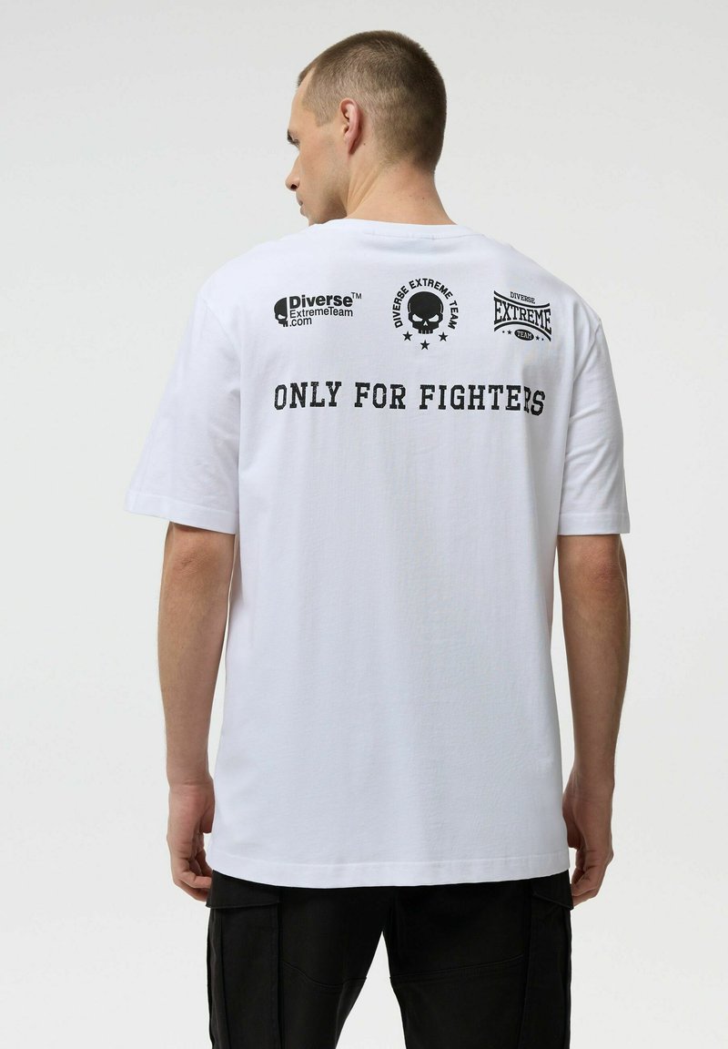 Mężczyzna w białej koszulce z napisem "ONLY FOR FIGHTERS" i logo Diverse Extreme Team na górnej części pleców, w połączeniu z czarnymi spodniami.