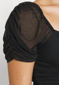 Haut noir avec des manches bouffantes transparentes et des détails froncés, accompagné d'un corps ajusté. Le tissu présente une finition texturée et un léger éclat.