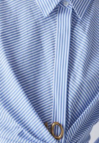 Camicia a righe blu e bianche con colletto, caratterizzata da un accento di anello dorato e una superficie in tessuto liscia. I design includono righe verticali.
