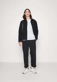 Casaco preto de botões por cima de uma t-shirt branca, combinado com calças pretas e ténis claros, apoiado contra um fundo branco liso.