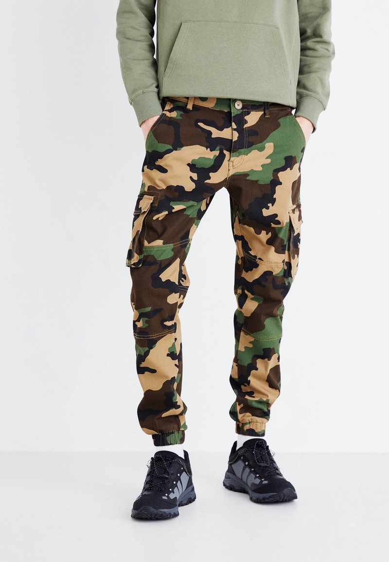 Persoon in een groene hoodie, camouflage cargobroek met zijzakken, witte sokken en zwarte wandelschoenen, staand met de handen in de zakken.