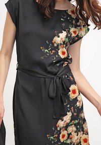 Černé květinové šaty z lehké tkaniny; mají měkké krátké rukávy, pásek s vázáním v oblasti pasu a živý květinový vzor v oranžové a zelené barvě.
