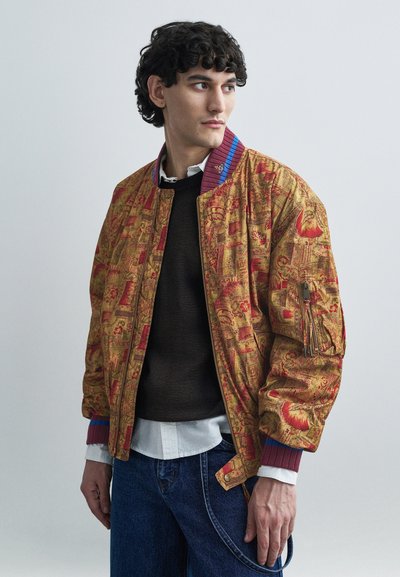 Bomber con motivo a fantasia in oro e rosso con collo a costine blu e rosso. Presenta tasche laterali e tessuto strutturato. Indossato sopra un maglione scuro.