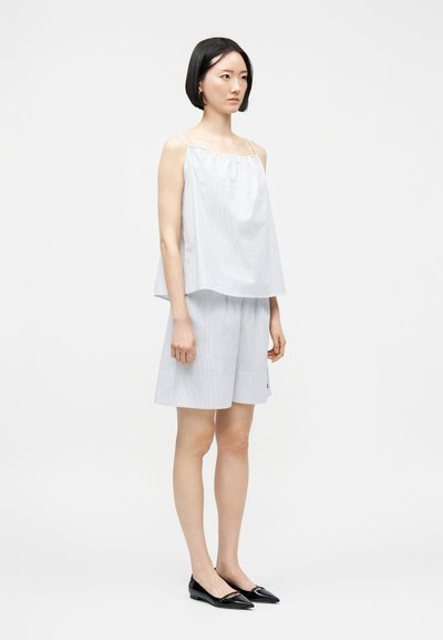 Γυναίκα με κοντά μαύρα μαλλιά φοράει λευκό αμάνικο τοπ και λευκά σορτς μέχρι το γόνατο, συνδυασμένα με μαύρα μυτερά παπούτσια, στέκεται πλαγίως.