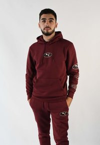 Conjunto de sudadera y pantalones de chándal color granate. La sudadera cuenta con cordones, un bolsillo canguro y parches con logo en el pecho y las mangas. Textura de tela suave.