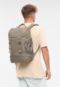 Harmaa kangasreppu, jossa on sileä koostumus, säädettävät olkahihnat, sivuilla verkkotasku ja vetoketjullinen pääosasto.