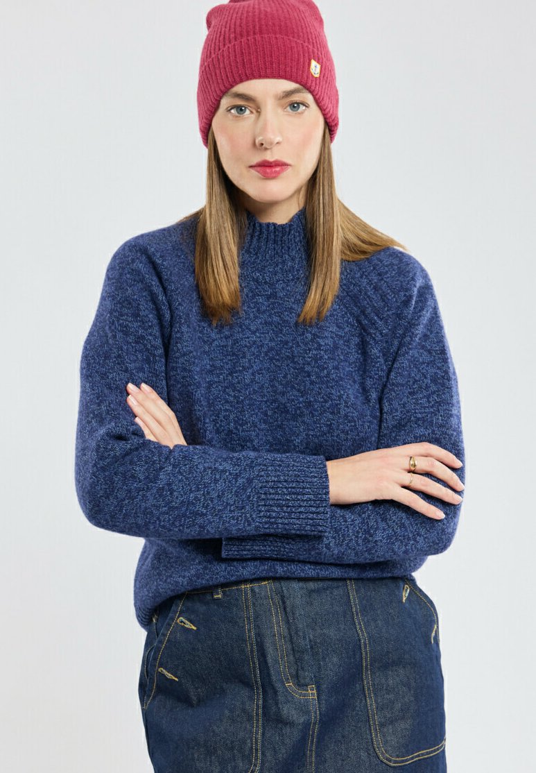 Pull en maille bleu marine avec un col montant et des poignets côtelés, associé à un bonnet côtelé bordeaux. La tenue est complétée par une jupe en denim.