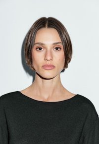 Una donna con capelli corti e lisci e un trucco neutro fissa direttamente lo spettatore, indossando un maglione scuro e largo su uno sfondo chiaro.