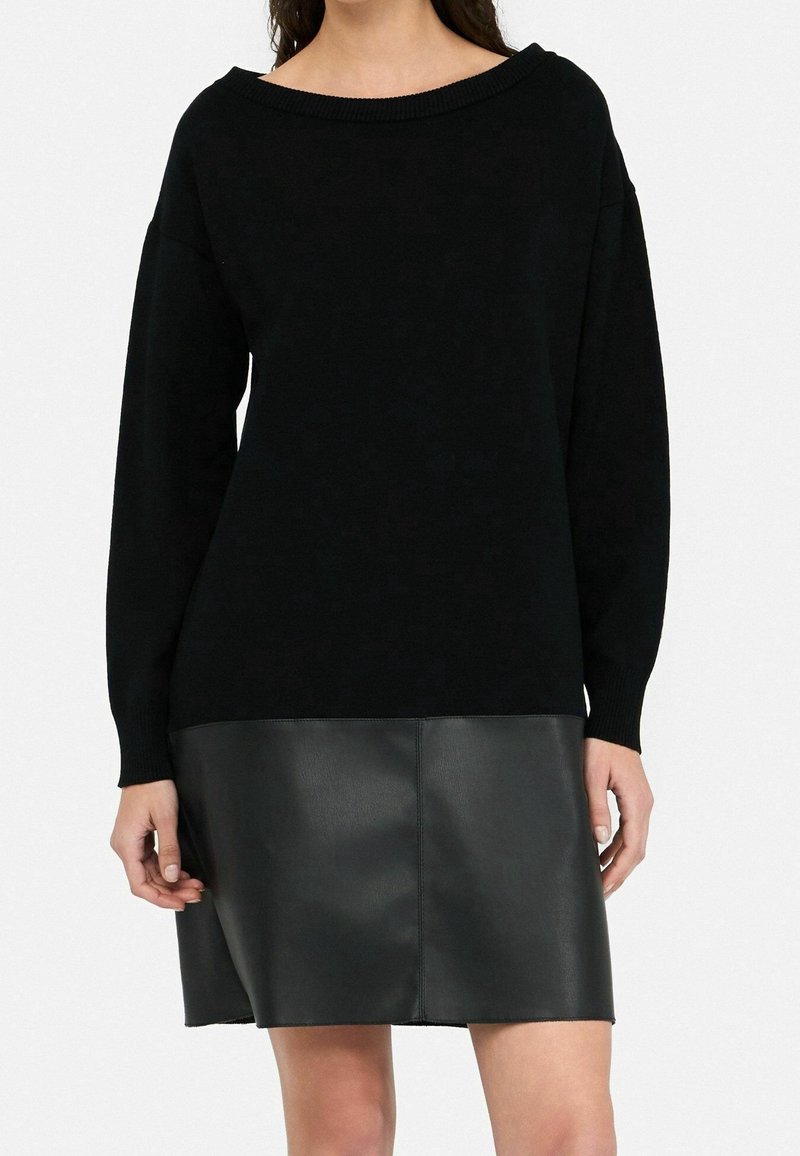 Femme portant une robe-pull noire à manches longues avec un haut en tricot et une jupe en cuir synthétique, debout devant un fond uni.