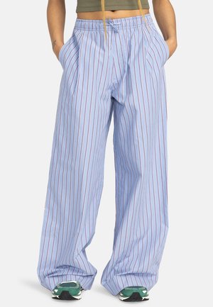 Pantaloni larghi azzurri chiari con righe rosse verticali, vita elasticizzata con coulisse, indossati con sneaker verdi e mani nelle tasche.
