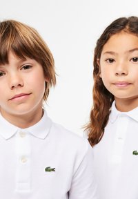 Due bambini indossano magliette polo bianche con piccoli loghi di coccodrillo verdi. Il tessuto ha un aspetto texturizzato con un design classico del colletto.