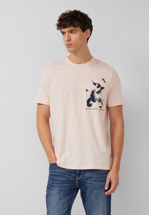 Jonge man die een lichtroze T-shirt draagt met een zwarte abstracte print en tekst, gecombineerd met een blauwe spijkerbroek, staand tegen een effen grijze achtergrond.